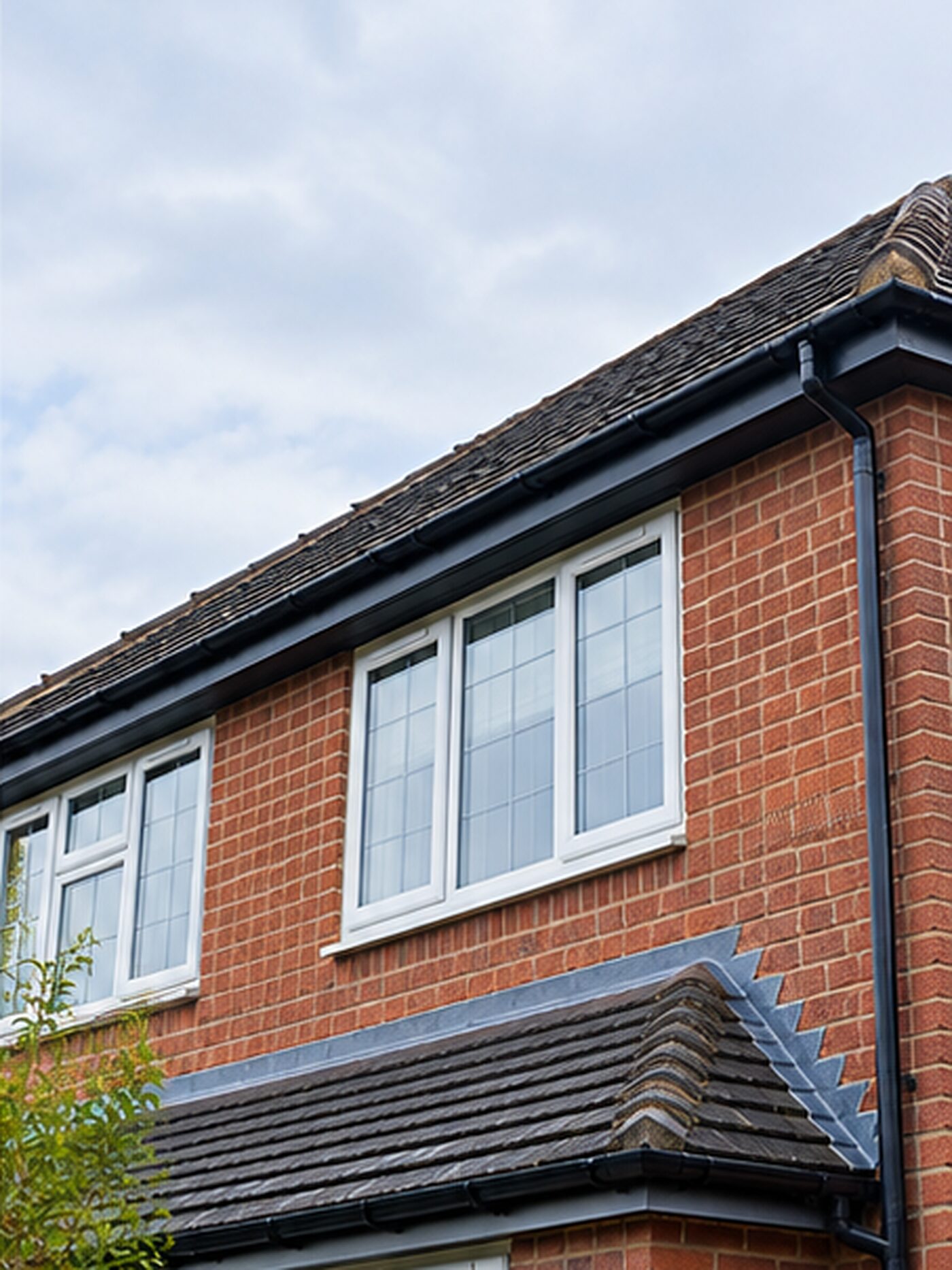 The roofline of a red-brick British house with clean black guttering and downpipe against an overcast sky